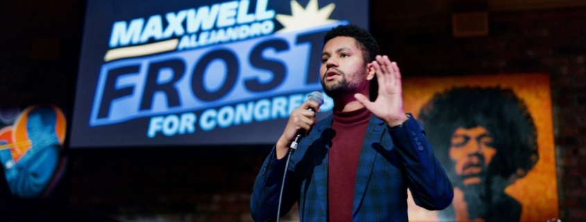 Maxwell Frost speaking on stage in a blue suit, gesturing with left hand