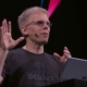 John Carmack speaking in front of a lectern with black and pink background behind