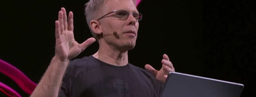 John Carmack speaking in front of a lectern with black and pink background behind