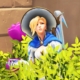 Mercy from Overwatch hiding in a bush. She is shrugging. She seems confused.