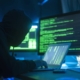 A stylised photograph of a person acting as a hacker, break into servers and infecting them with a virus, as show by computer monitors displaying green text and codes