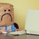 A child with a sad paper bag on their head sits staring at an old PC.