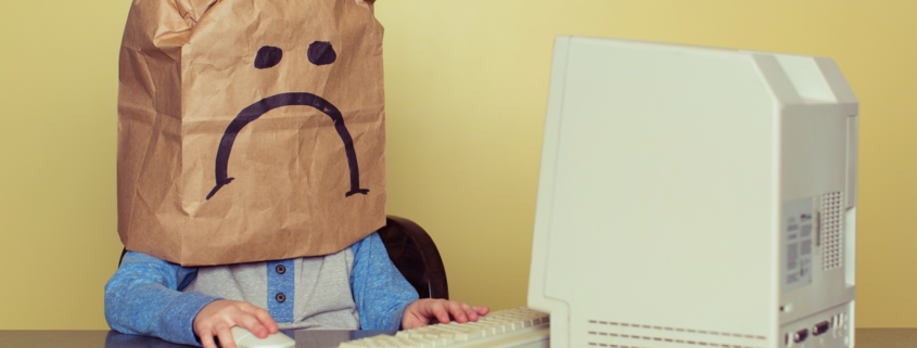 A child with a sad paper bag on their head sits staring at an old PC.