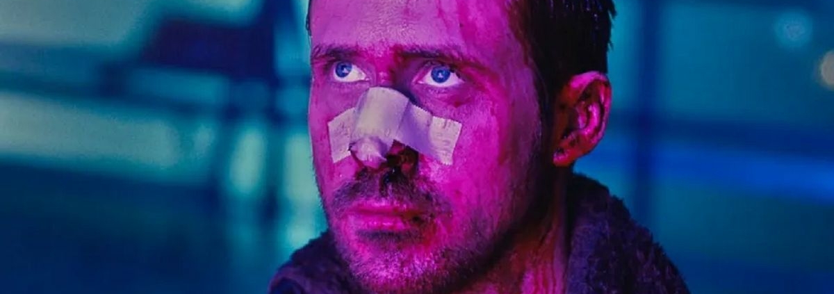 Ryan Gosling in Blade Runner: 2049, his face cut up and with a bandage over his nose, bathed in purple light with the blackground a blurry blue